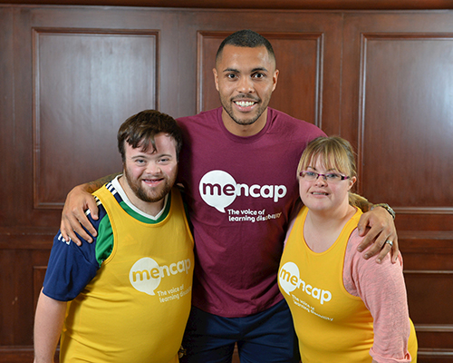Josh Magennis smiling wearing a pink Mencap t-shirt stood with his arms around two people who are wearing yellow Mencap running vests