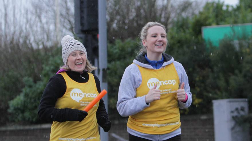 Two women smiling whilst running together, they are wearing yellow Mencap running vests and one is holding a relay baton.