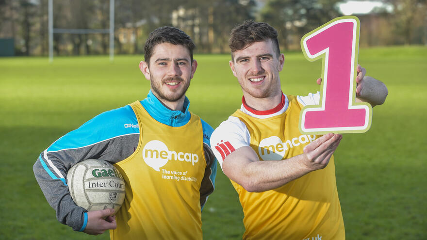 Two men stood together on a football pitch, one holding a sign the other holding a football