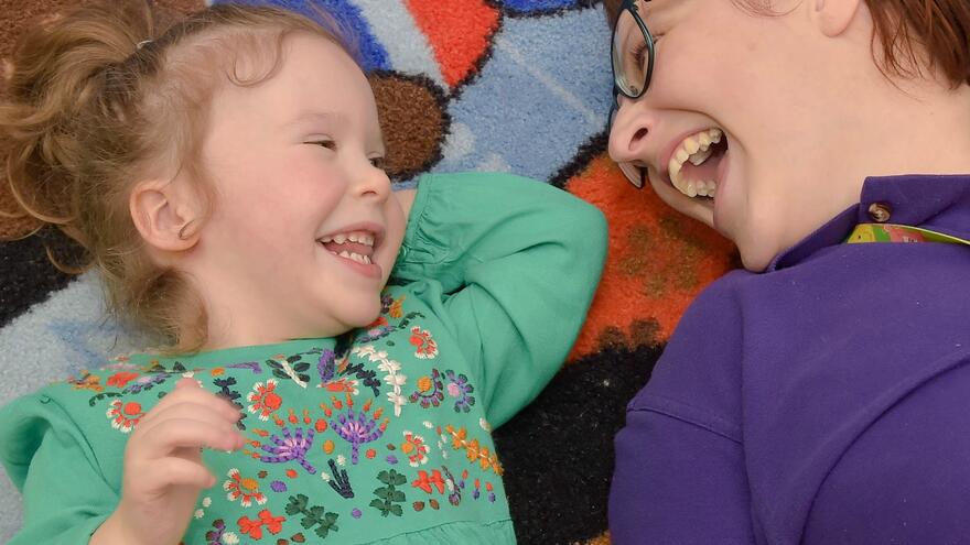 Woman lying down next to young girl. They are both laughing and looking at each other.