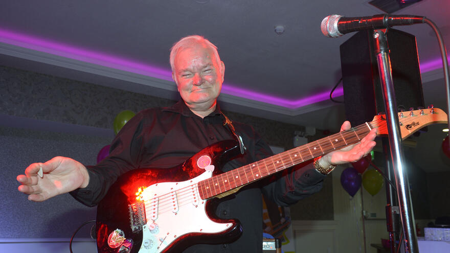 Older gentleman on stage wearing a black shirt playing a guitar in front of a microphone.