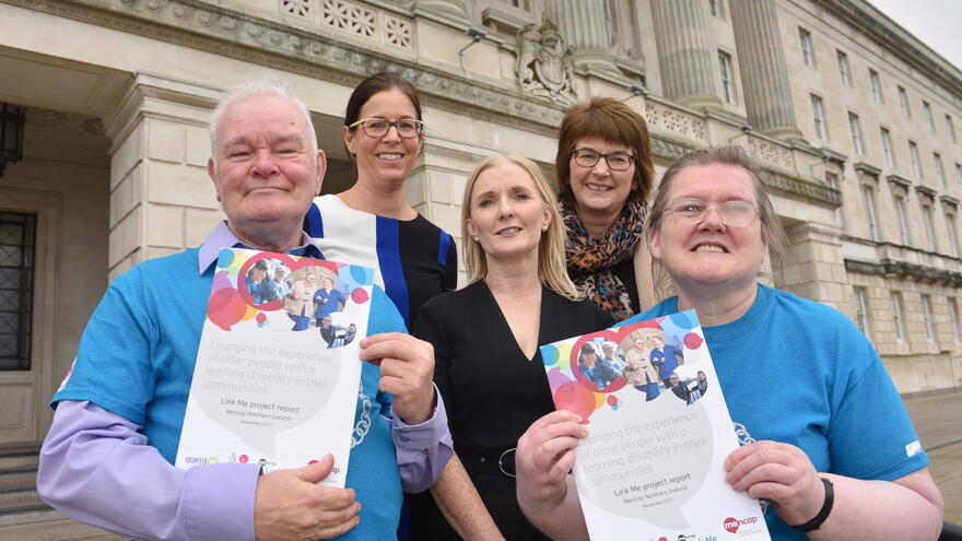 Group of 5 people stood outside building smiling. Two people are holding copies of the Link Me project report.