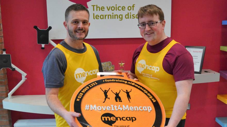 Two men stood inside in front of a Mencap sign, wearing yellow Mencap running vests, each holding on to a sign that reads #MoveIt4Mencap