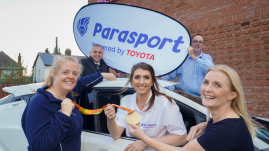 People stood together around a car, people in the background holding a "Parasport sponsored by Toyota" sign