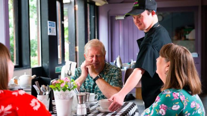 A waiter with downs syndrome is serving three people at a table in a cafe with a cup of coffee