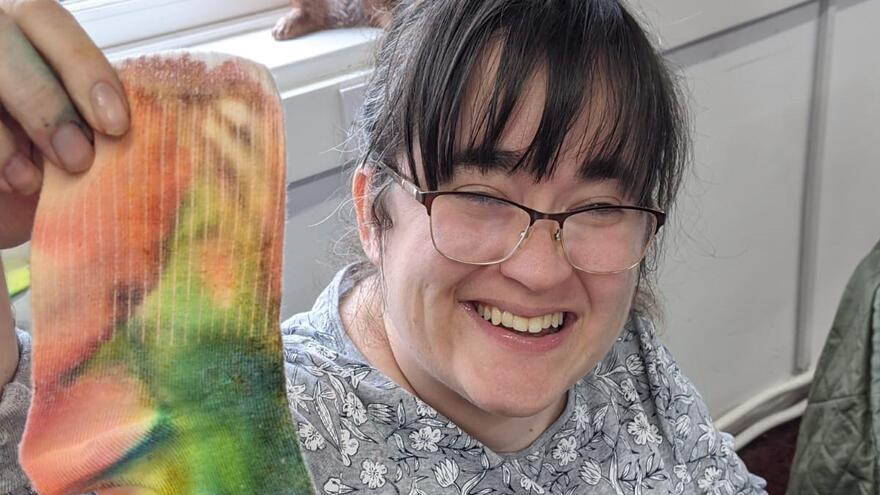 A smiling woman sitting on a chair holding up a colourful tie dyed sock