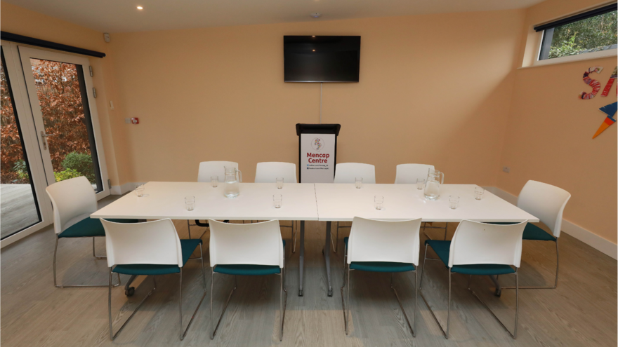 Image of long table with chairs set up surrounding the table. Behind the table is a lectern, above that on the wall is a TV which is off.