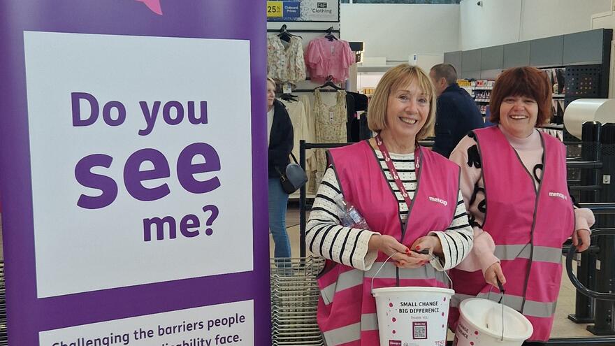 Two volunteers wearing Mencap hi-vis vest standing with buckets in Tesco