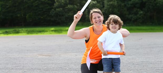 Woman wearing running kit knelt down outside next to her young son. They are both holding relay batons.