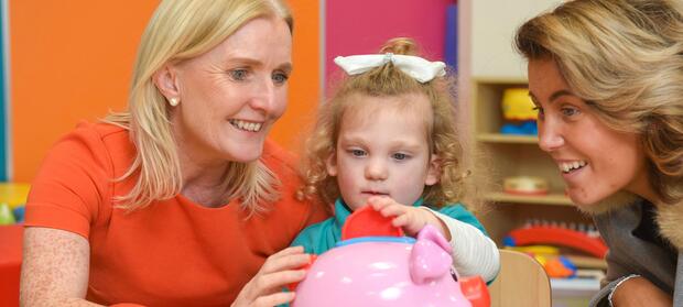 Two women sat in nursery with young girl in between them who is playing with a pink toy.