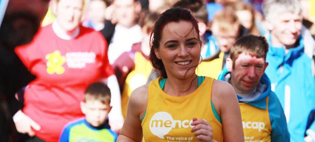 Female runner wearing yellow Mencap NI running vest smiling whilst running in crowd of runners.