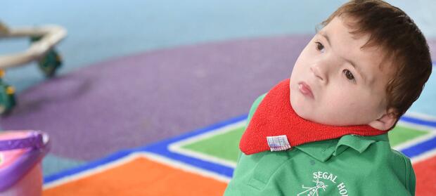 Young boy with brown hair, wearing red bib and green polo shirt sat on colourful floor tiles