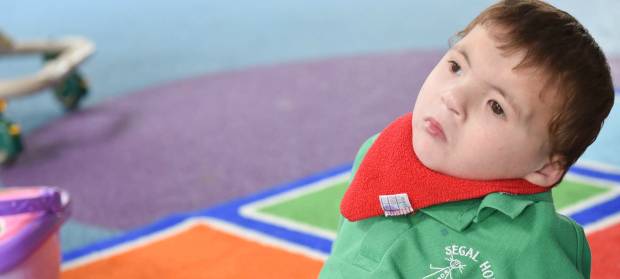 Young boy with brown hair, wearing red bib and green polo shirt sat on colourful floor tiles