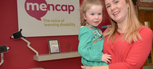 Mother holding her daughter in front of Mencap sign.