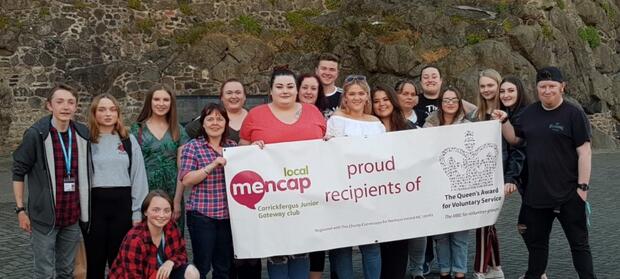 Group of people stood together holding large banner which reads "Proud recipients of the Queen's Award for Voluntary Service"