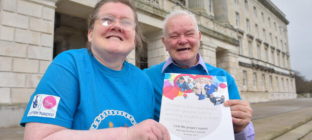Two people stood smiling outside building holding booklet.